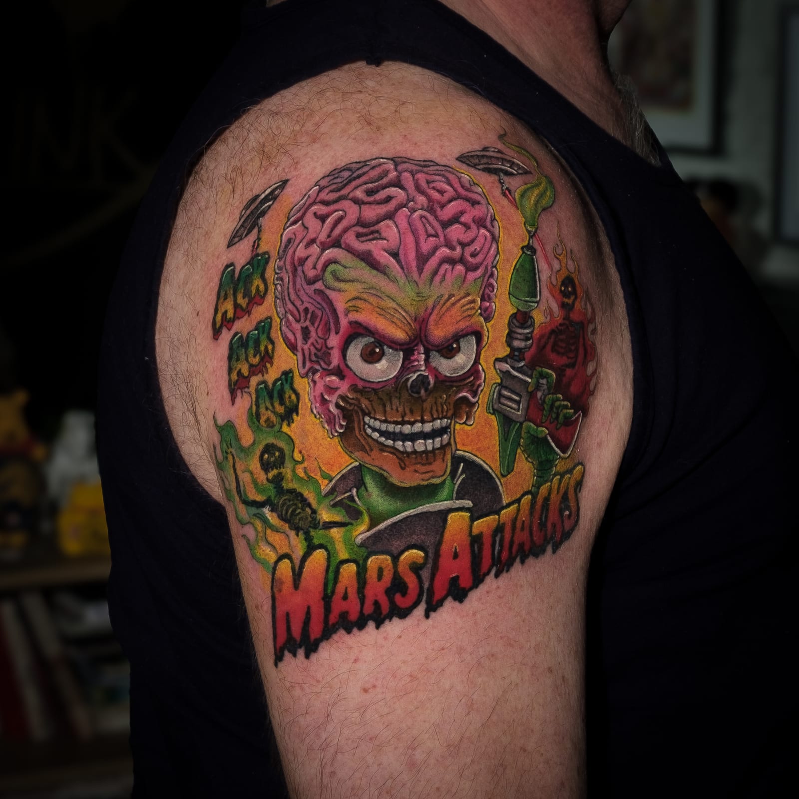 Colour Mars Attacks tattoo of an alien with an exposed brain and ray guns on an upper arm.