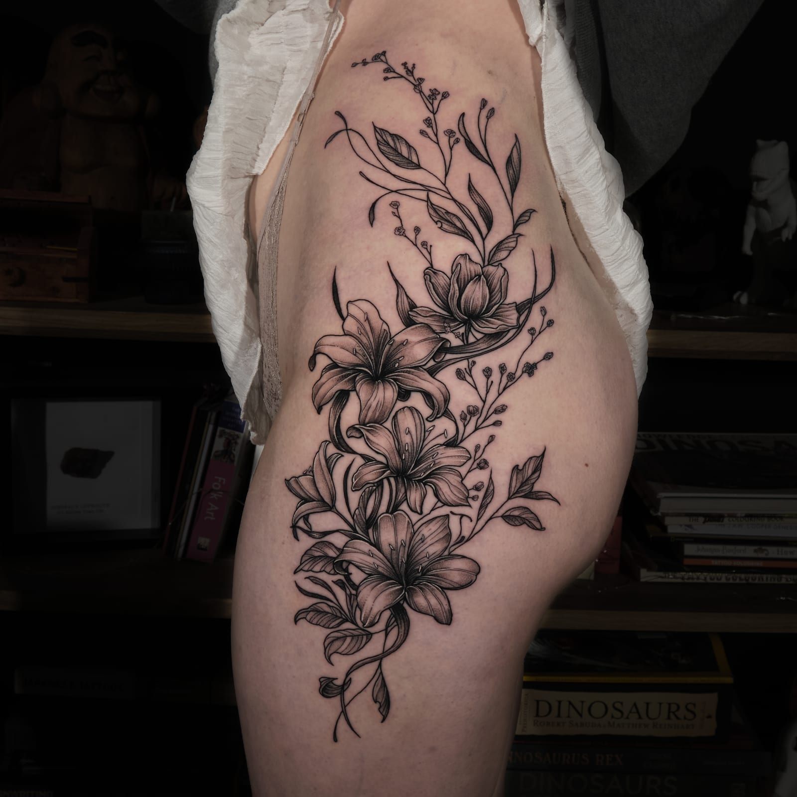 Fine-line floral tattoo of lilies and botanical stems flowing along the side of the hip and thigh.