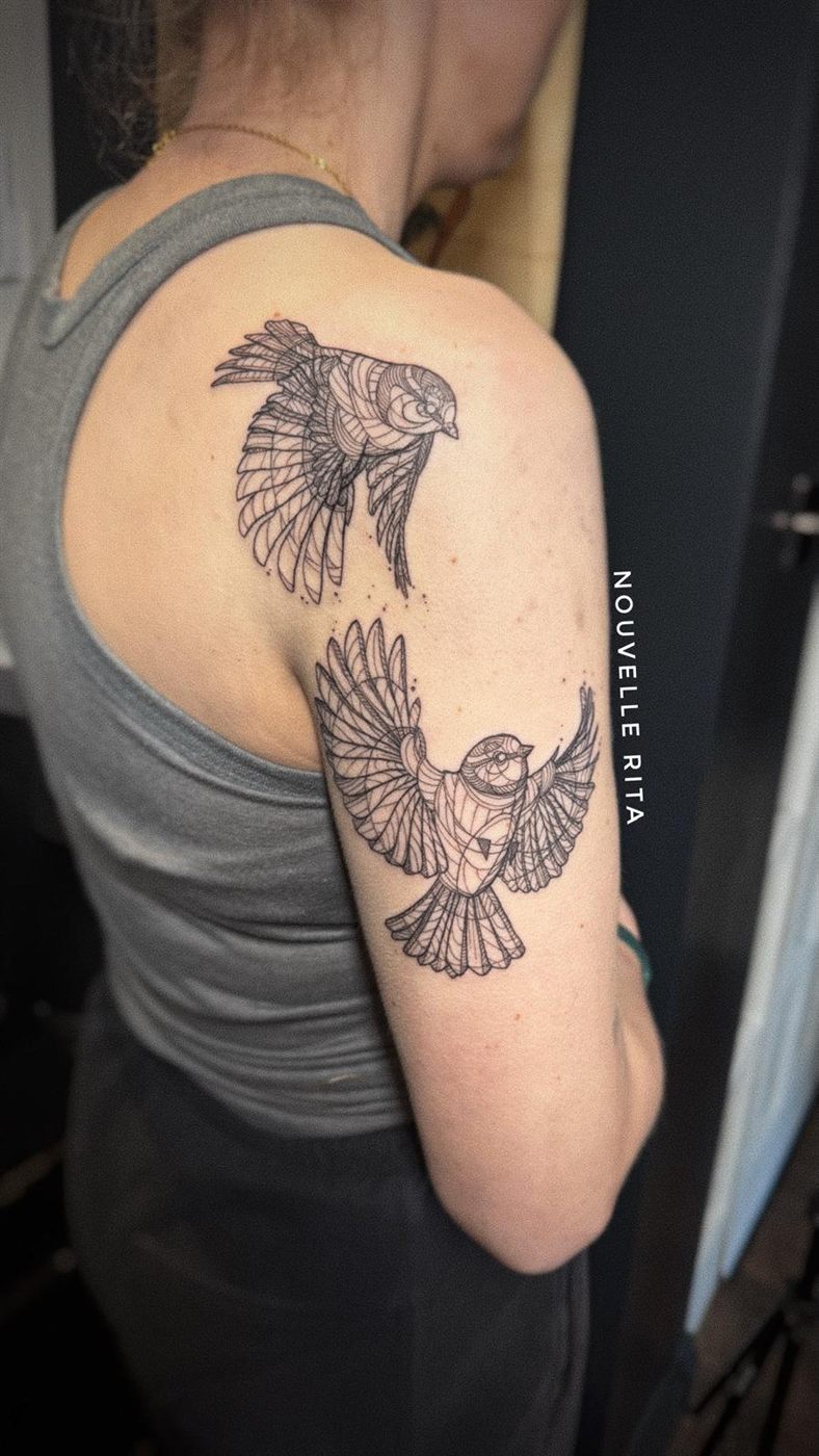 Fine-line tattoo of two small birds in flight across a shoulder and upper arm.